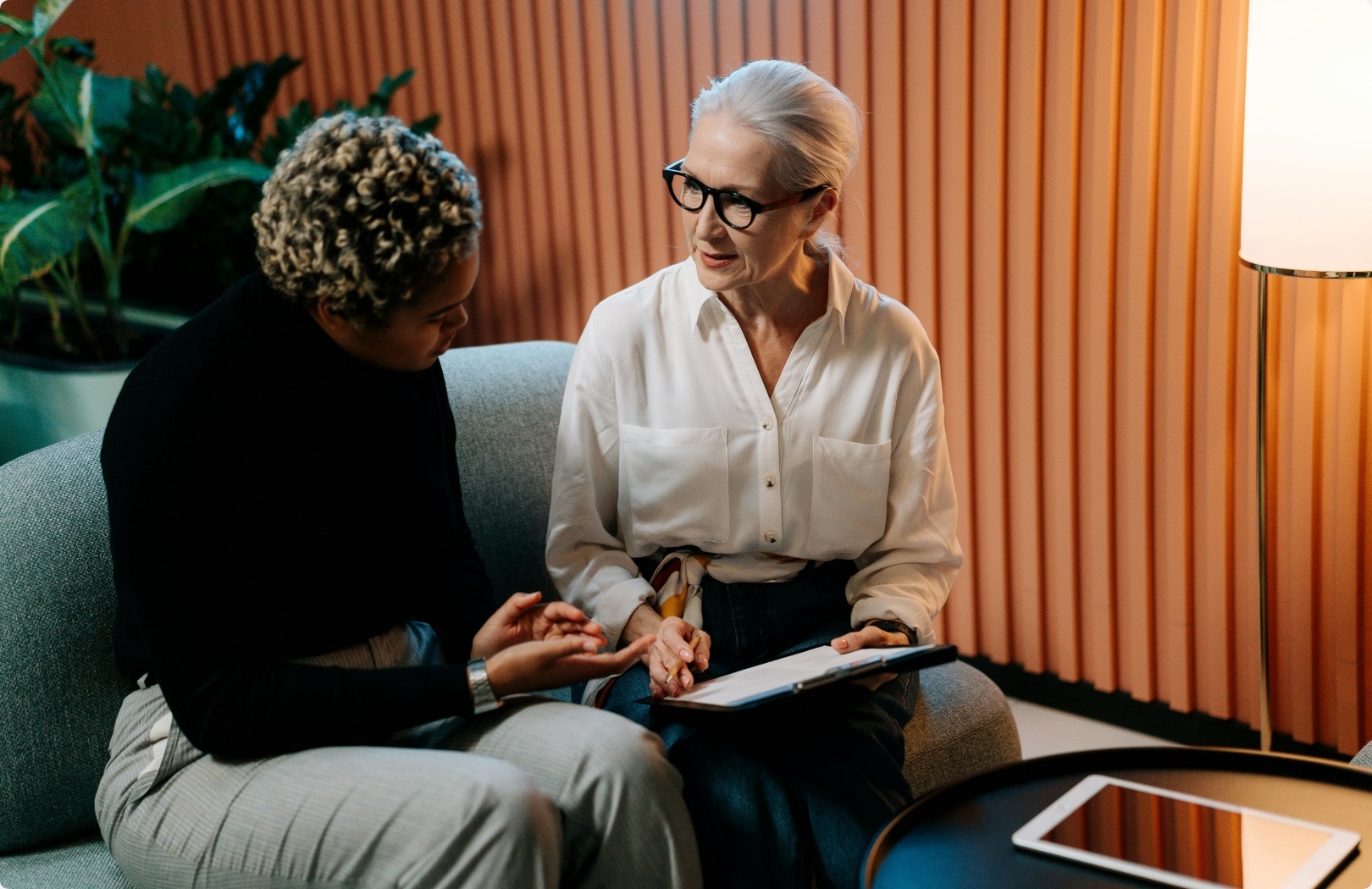 Two women are sitting on a sofa and having a conversation. An older woman with gray hair and glasses is holding a notebook, while the younger woman is listening.