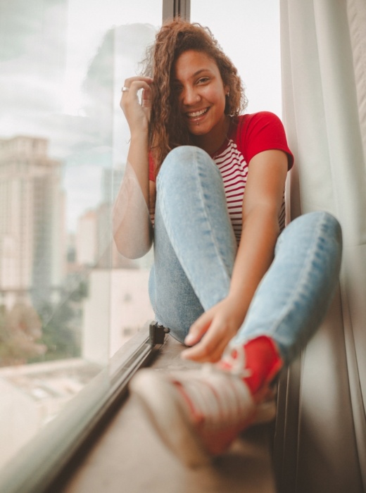 Eine lächelnde Frau sitzt in lässiger Kleidung am Fenster