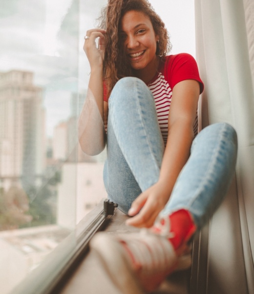 Eine lächelnde Frau sitzt in lässiger Kleidung am Fenster