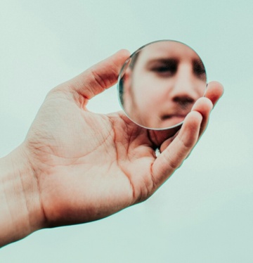 Hand, die einen runden Spiegel hält, in dem das Gesicht einer Person reflektiert wird.