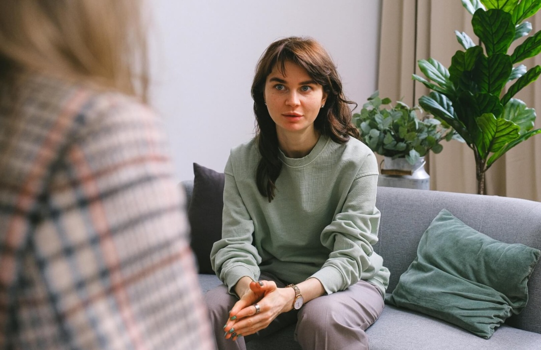 Frau in einem grünen Pullover sitzt auf einem Sofa und spricht mit einer anderen Person.