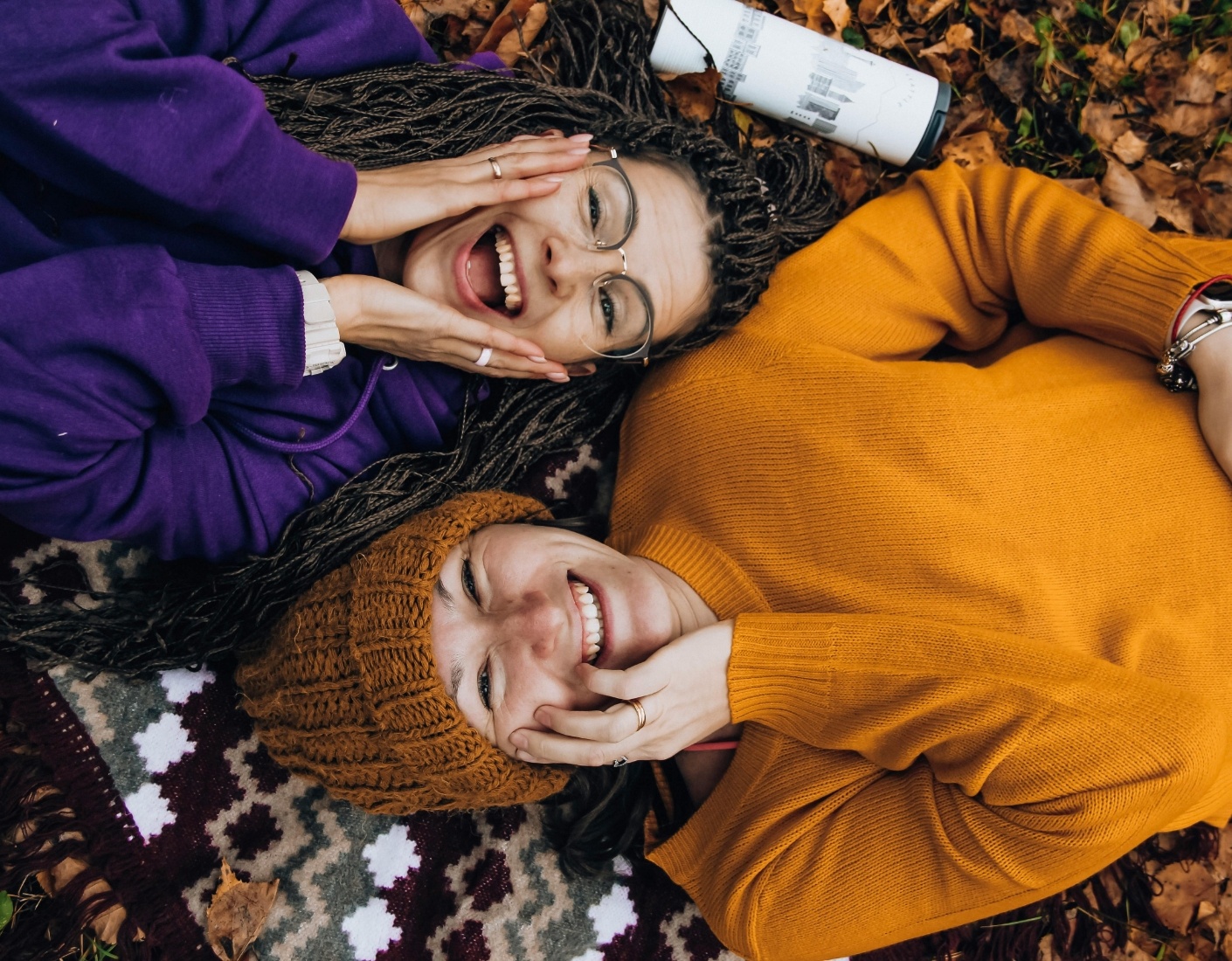 Two women lying on a blanket outdoors, both smiling and with their hands on their faces.