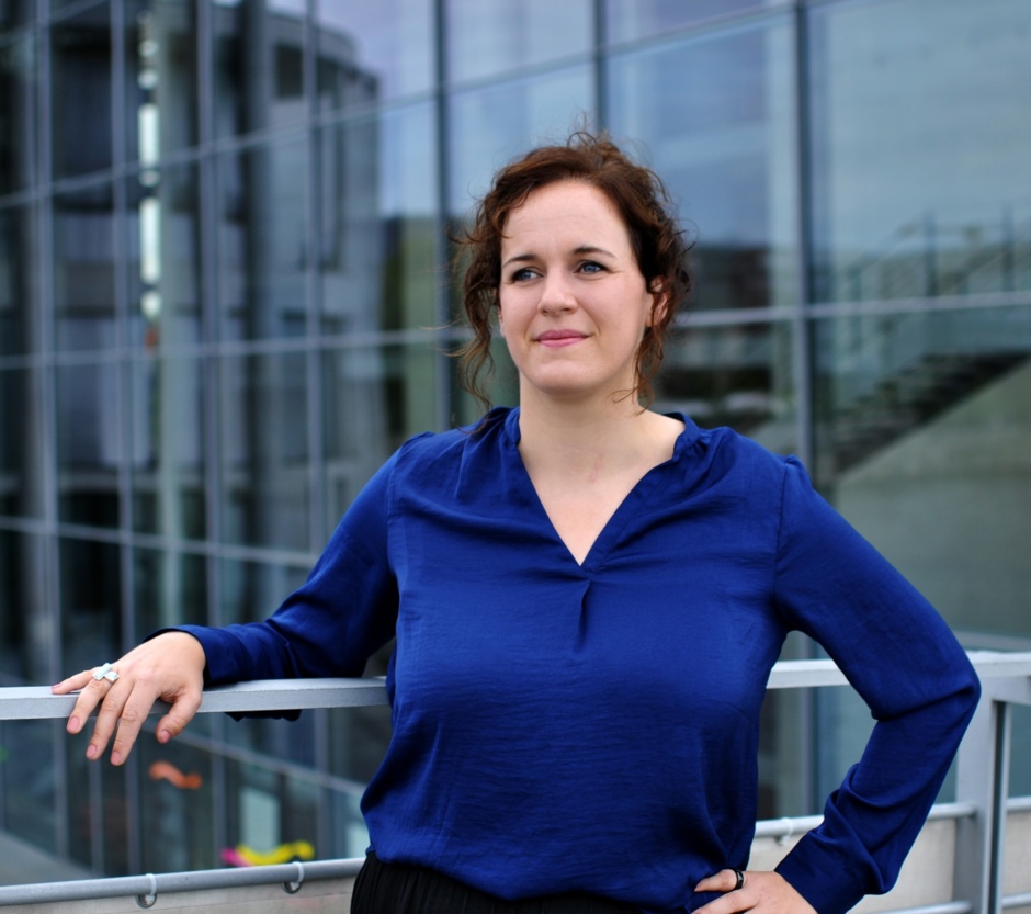 Katrin Terwiel steht in einer blauen Bluse vor einem modernen Gebäude und hält ihre rechte Hand auf einem Geländer