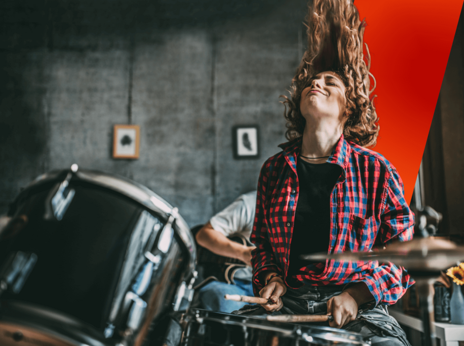 A woman with long hair passionately playing drums in a rehearsal room.