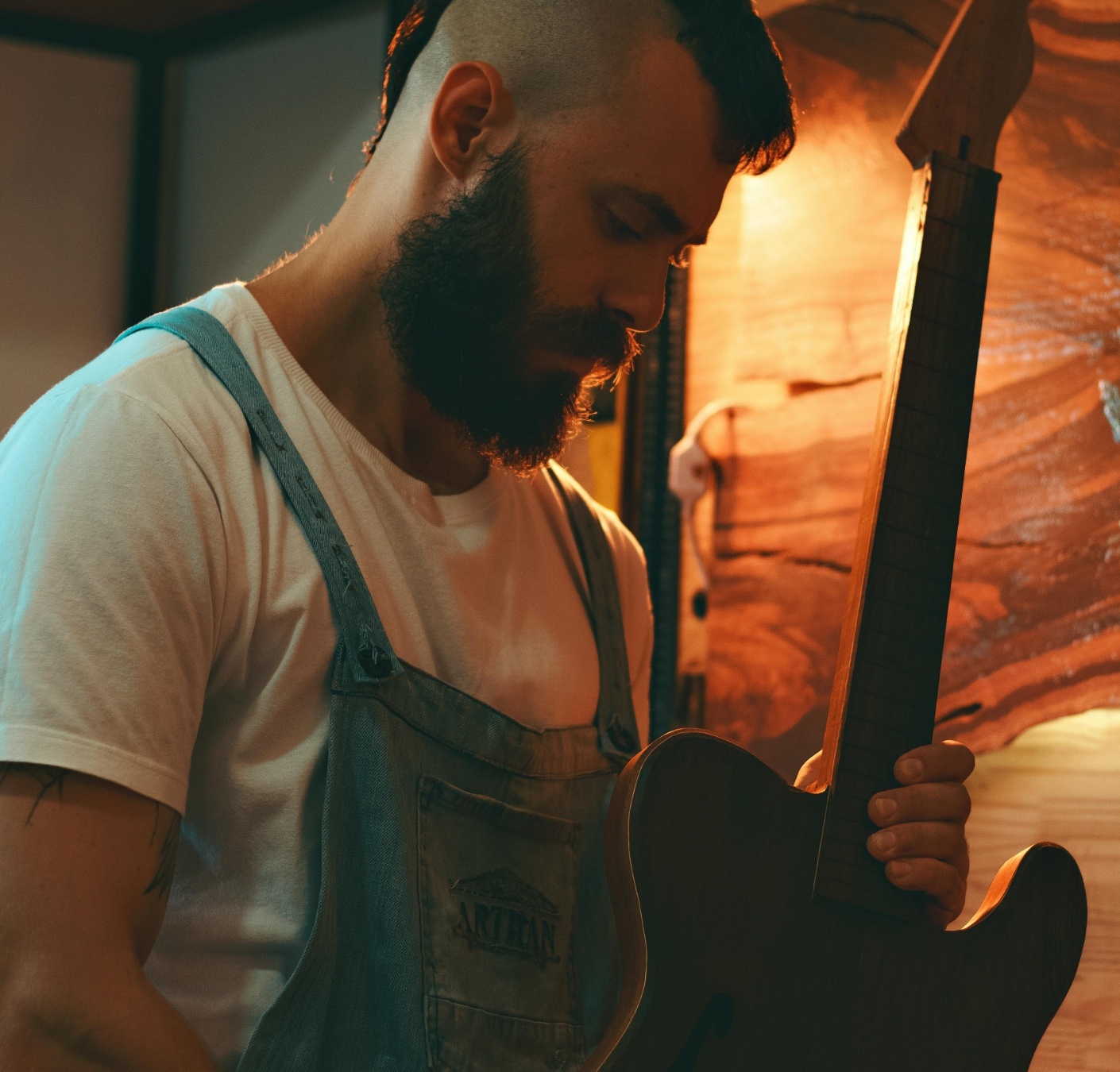 A man with a beard and mohawk hairstyle working on a guitar in a workshop.