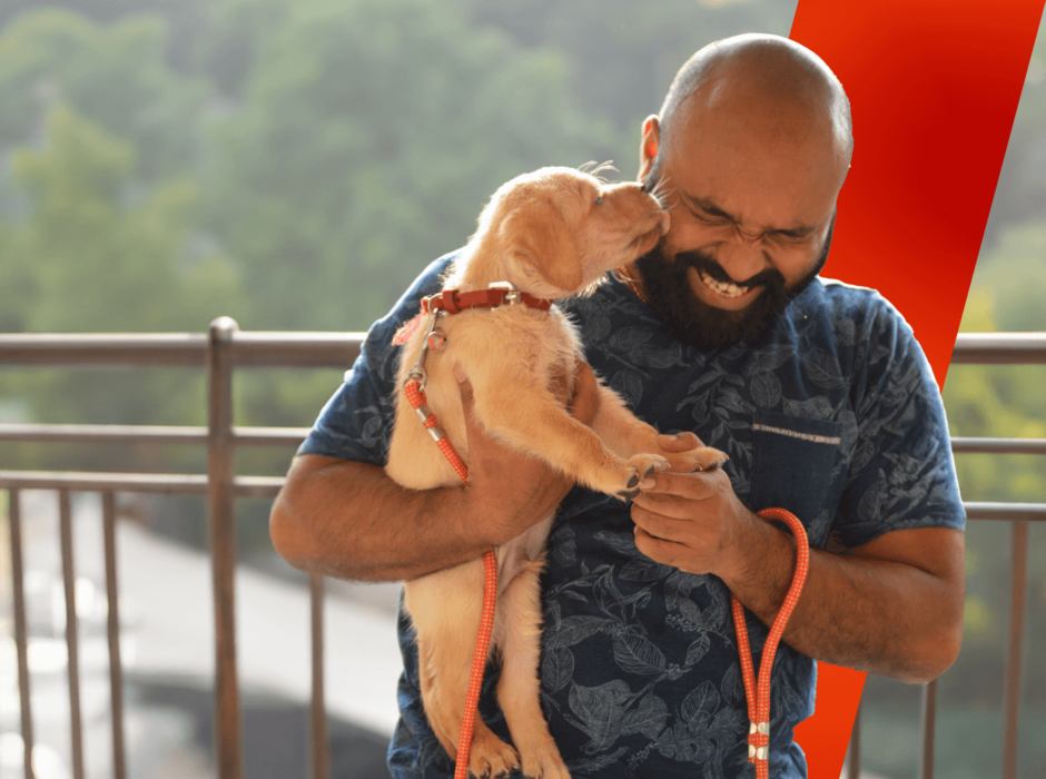 A laughing man with a beard holds a playful puppy licking his face, outdoors.